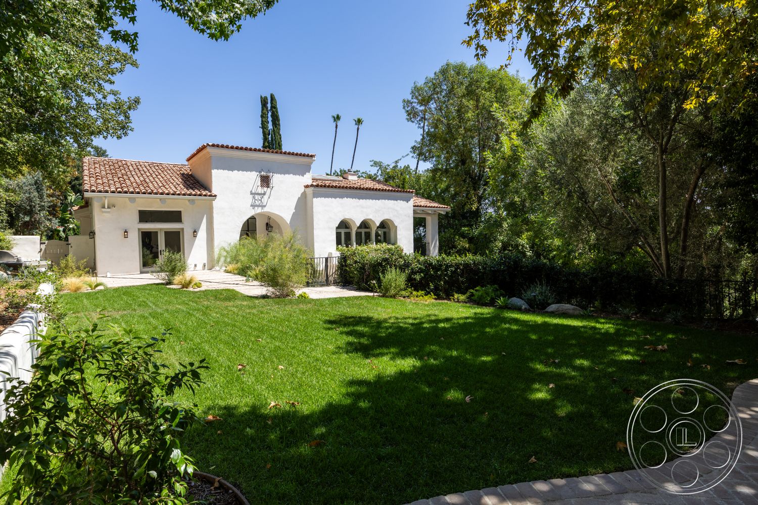 Medit 168 - exterior, landscaped garden, white stucco walls, potted plants, circular driveway, stone pathway, lawn area, green shrubbery, terracotta tile roof, arched windows, tall palm trees, suburban backyard, wooden front door, Mediterranean architectural style, decorative wall lanterns, gallery covered porch