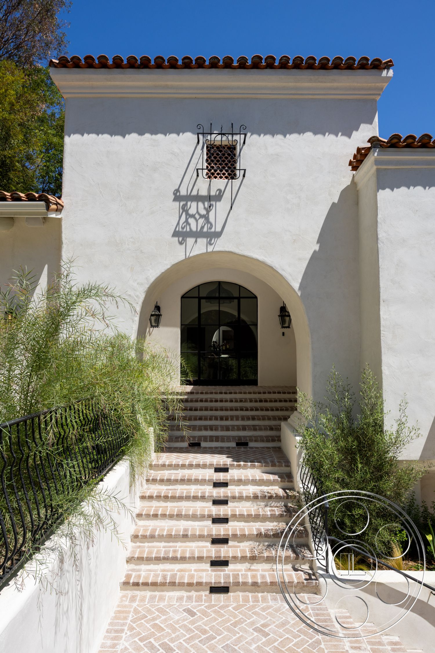 Medit 168 - exterior, terracotta roof tiles, stucco walls, arched entryway, cobblestone path, Mediterranean architectural style, black metal railings, textured plaster finish, landscaped garden areas, tall arched windows, herringbone brick steps, decorative wrought iron detailing, sun-drenched entryway