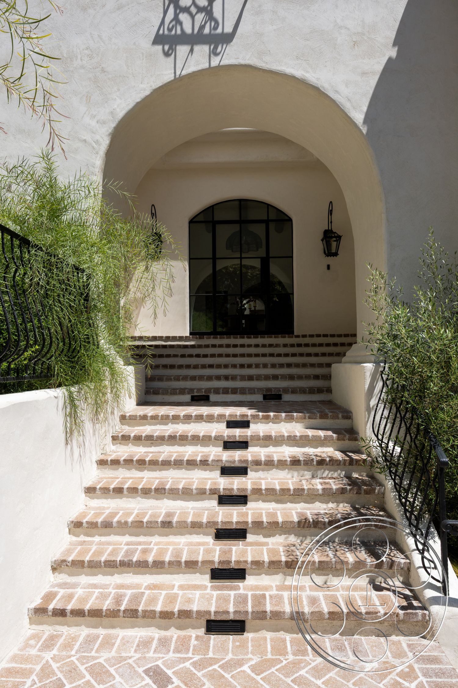 Medit 168 - exterior, landscaped garden, black metal railing, stucco walls, arched entryway, textured plaster wall, brick paver steps, black wall-mounted lantern, large arched window, integrated step lighting, cobblestone paver driveway