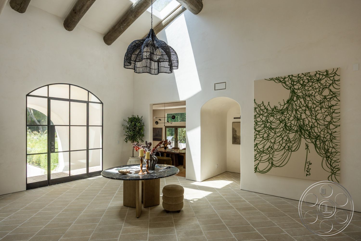 Medit 168 - woven pendant light, arched doorways, textured plaster walls, round marble dining table, stone tile flooring, vaulted ceiling with exposed wood beams, colorful abstract wall art, large glass entry door, window views of green landscape