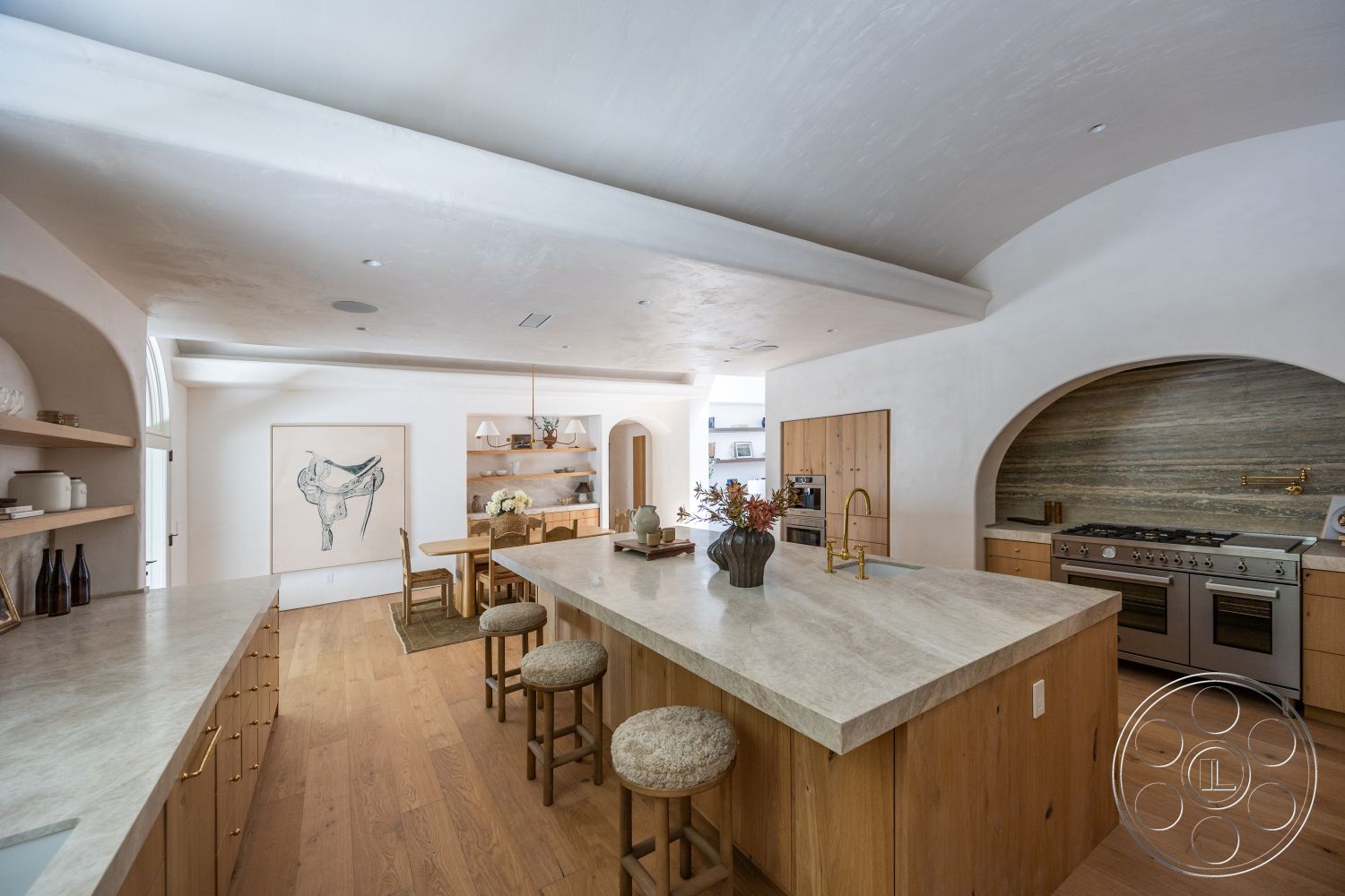 Medit 168 - minimalist decor, neutral color palette, stainless steel appliances, natural wood flooring, light wood cabinetry, decorative wall art, textured plaster ceiling, glass light fixtures, built-in cabinetry, open-concept kitchen, large island with stone countertop, waterfall edge countertop, arched kitchen entryway, exposed wood shelves, gold-toned faucet, luminous ambiance