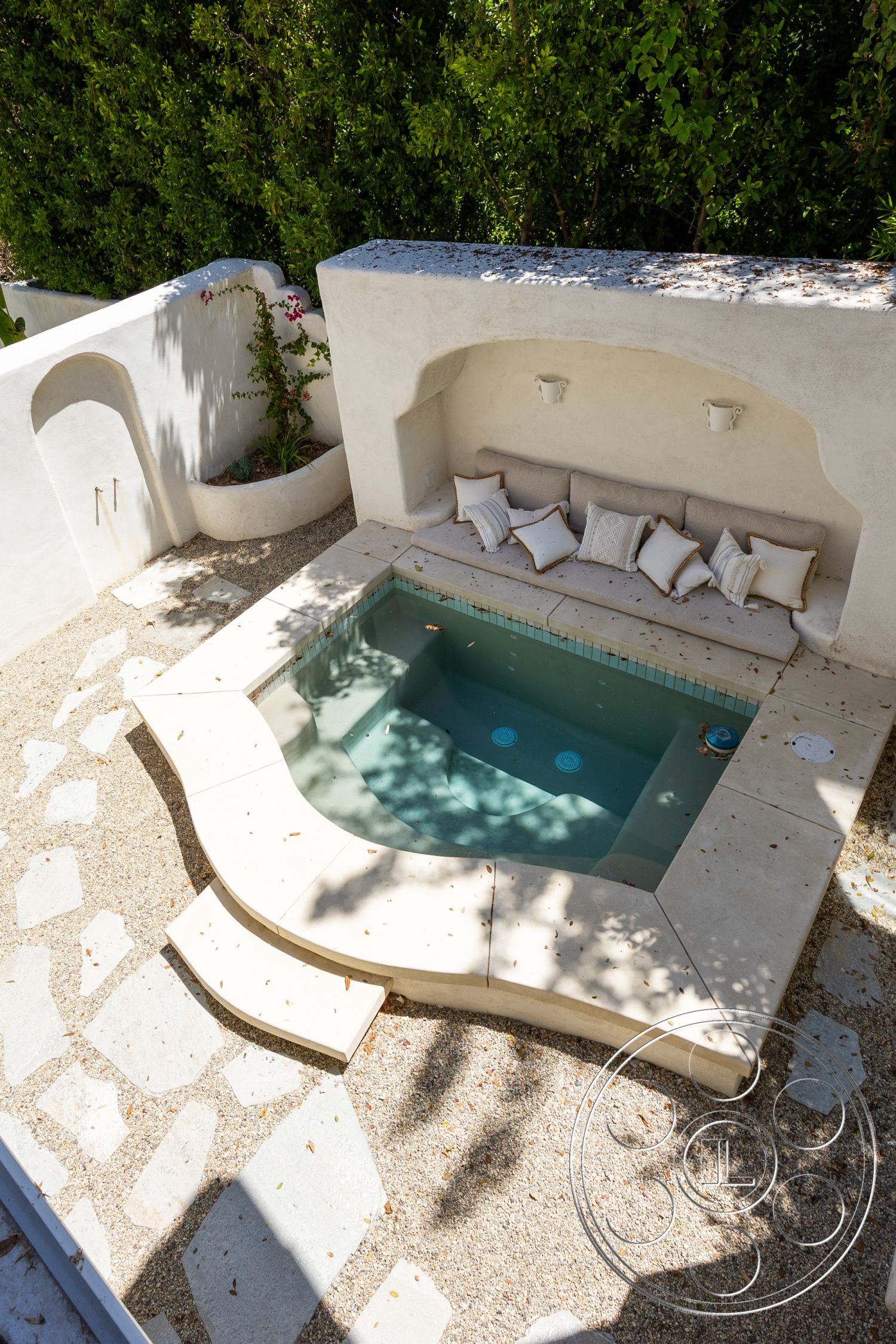Medit 168 - exterior, plaster wall finish, lush green landscaping, landscape lighting fixtures, natural stone patio flooring, smooth concrete coping, curved hot tub deck, rectangular hot tub, built-in seating area with soft cushions, decorative gravel surface
