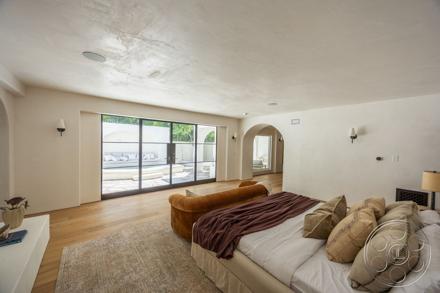 Medit 168 - minimalist decor, large glass sliding doors, open floor plan, textured ceiling, arched doorways, decorative throw pillows, modern upholstered bed, rectangular area rug, exposed plaster walls, light hardwood bedroom floors, sconce wall lights, sunlit room with outdoor view