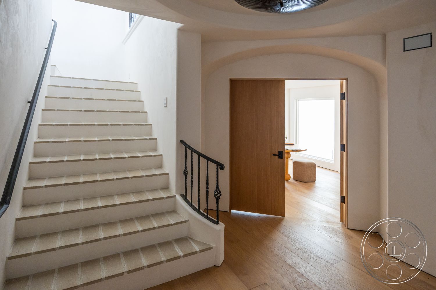 Medit 168 - light wood flooring, white plaster walls, natural light from large window, paneled wooden door, metal stair railings, modern ceiling light fixture, concrete stair treads, rounded archway entrance