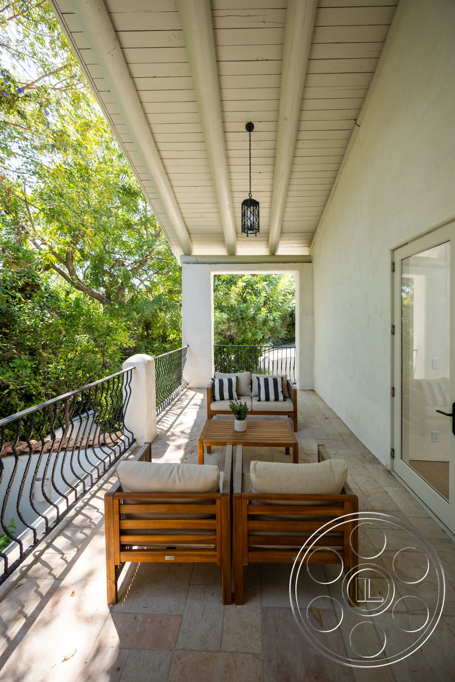 Medit 168 - exterior, white plaster walls, rectangular coffee table, wooden outdoor furniture, black wrought iron railing, striped outdoor cushions, metal pendant lighting, minimalist outdoor space, wood beam ceiling, lush greenery setting, open-air porch, stone patio flooring