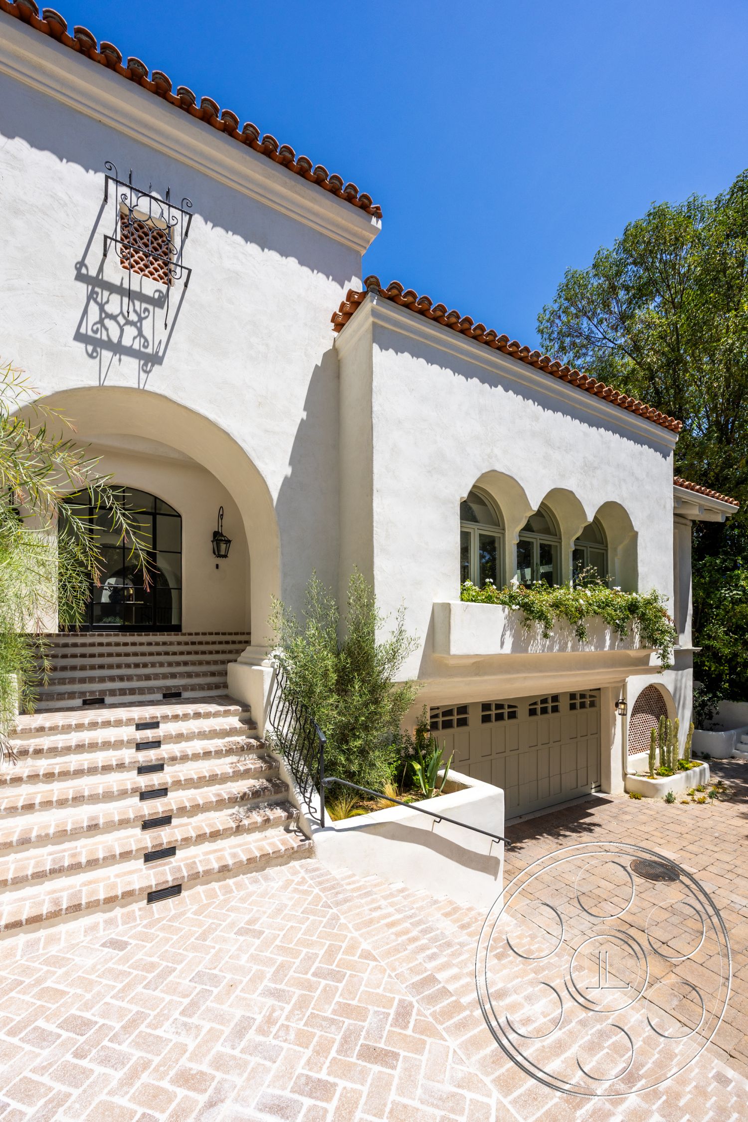 Medit 168 - exterior, urban setting, arched doorways, textured plaster walls, arched windows, clay tile roof, wrought iron railing, decorative wall lantern, brick driveway pavers, stucco house facade, landscaped garden with greenery, herringbone patterned driveway, concrete steps with integrated lighting, tiled entryway