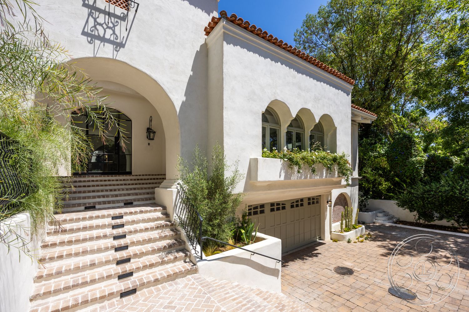 Medit 168 - exterior, landscaped garden, paver driveway, red tile roof, arched doorways, wrought iron railing, stucco exterior walls, lush greenery surrounding the property, arched window openings, brick steps with inlaid stone risers, terracotta flower boxes, rounded patio area, contemporary Mediterranean style, large front entry door, decorative lantern lighting