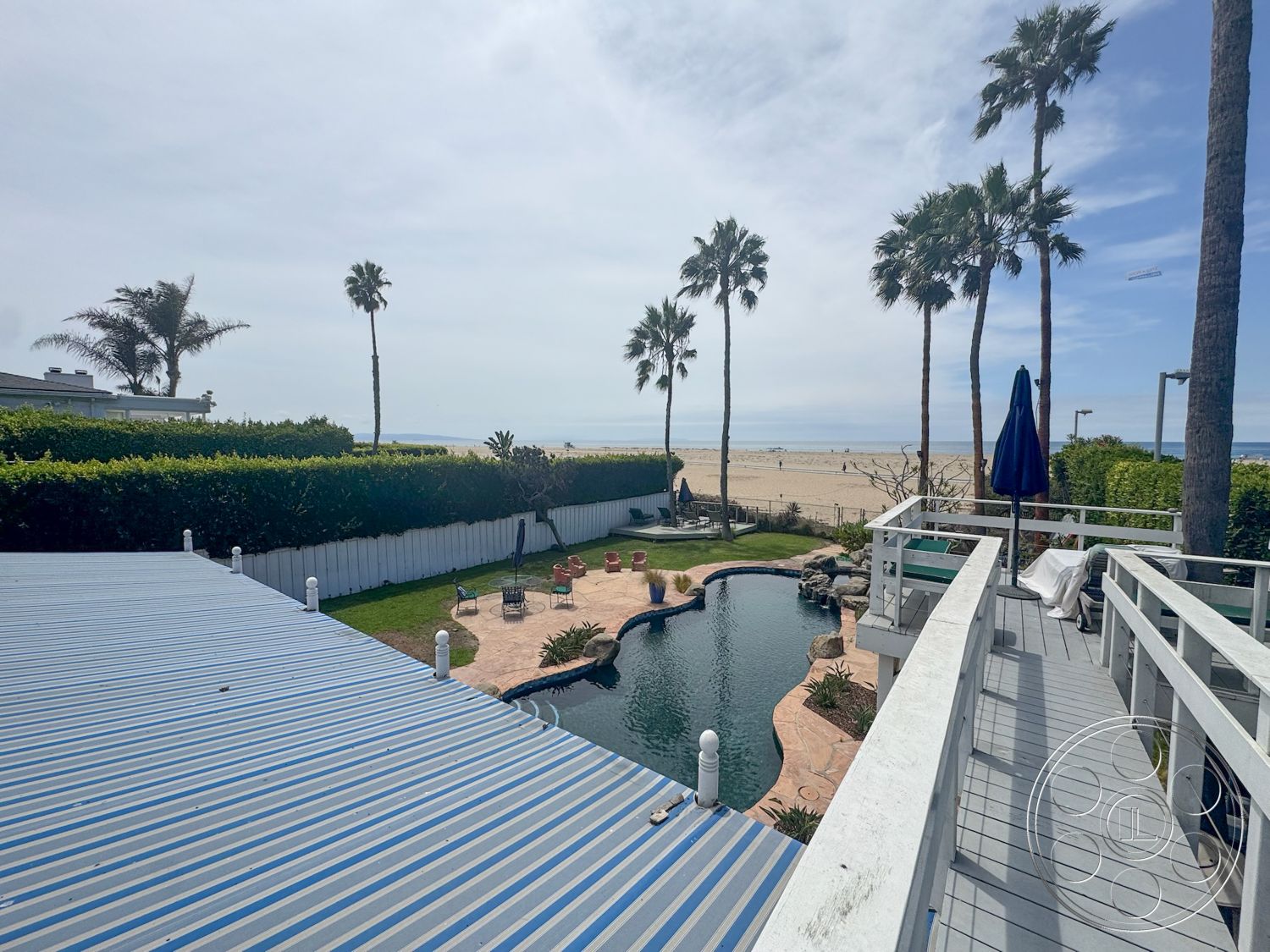 Lloyd Beach House - exterior, outdoor seating area, lawn area, white picket fence, tropical palm trees, striped awning, sunny outdoor space, paved pool deck, circular fire pit, curved swimming pool, landscaped garden area, concrete pool border, oceanfront view, wooden deck railing, textured patio flooring, beach setting