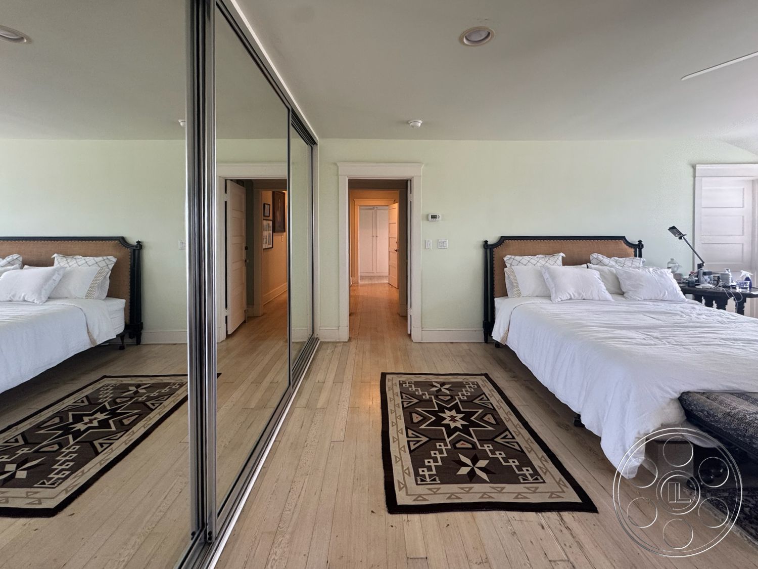 Lloyd Beach House - hardwood bedroom floors, crown molding details, textured area rug with geometric patterns, cream painted walls, queen-sized bed with decorative pillows, traditional wooden nightstand, mirrored sliding closet doors, white bedding with subtle patterns, doorway leading to hallway, decorative wall-mounted lamp, light-filled bedroom