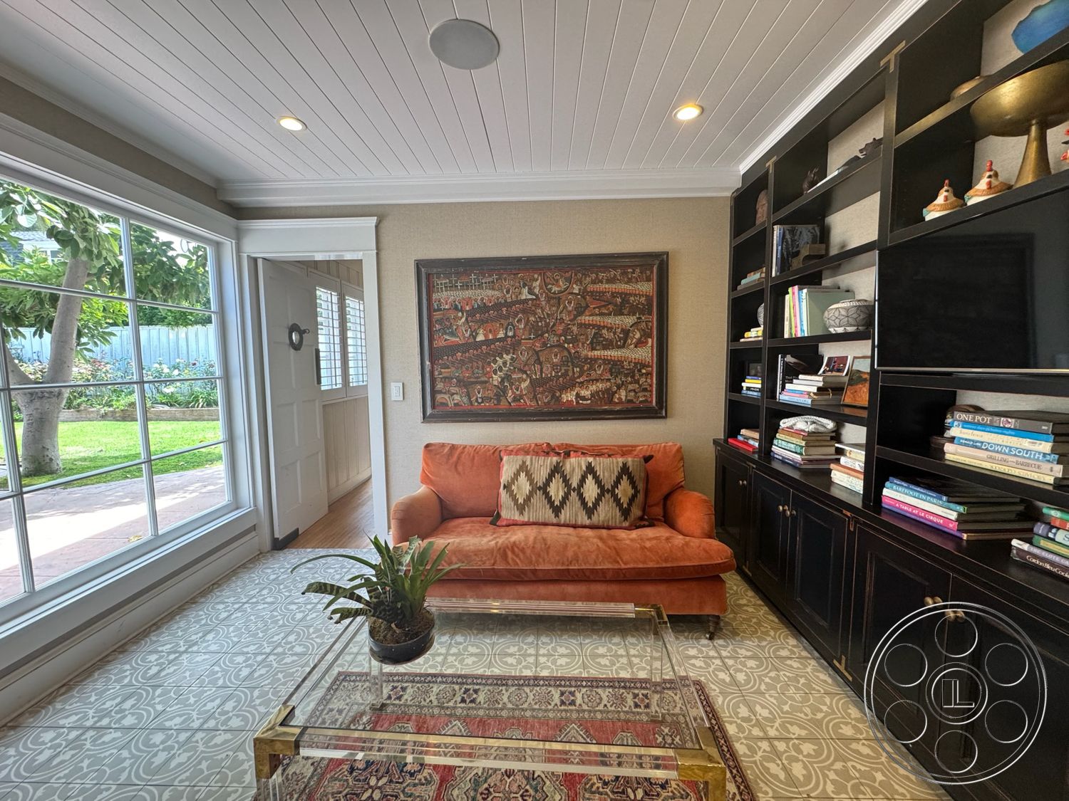 Lloyd Beach House - natural light, hardwood flooring, decorative area rug, glass coffee table, neutral wall color, framed wall art, green indoor plant, paneled door, vintage-style decor, upholstered orange sofa, textured fabric throw pillow, black wood bookshelves, open shelving with decorative items, large front window, white beadboard ceiling