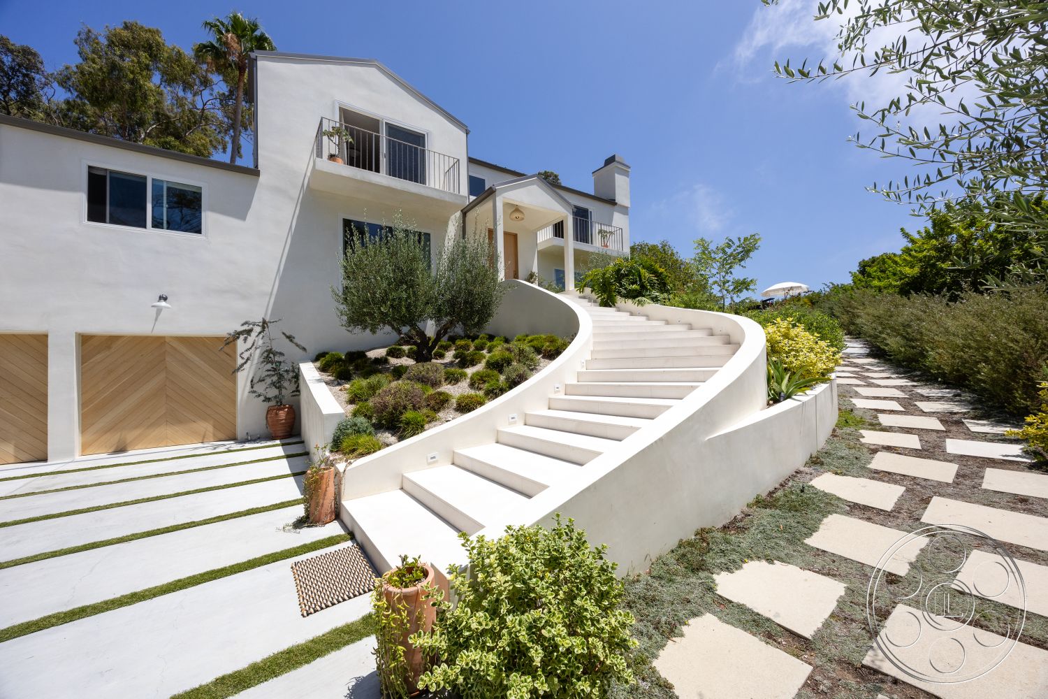 Americana 434 - exterior, stone pathway, modern architectural style, concrete driveway, stucco walls, landscaped garden area, planter pots with greenery, metal balcony railing, wooden garage doors, curved concrete stairs, lawn grass strips, split-level design