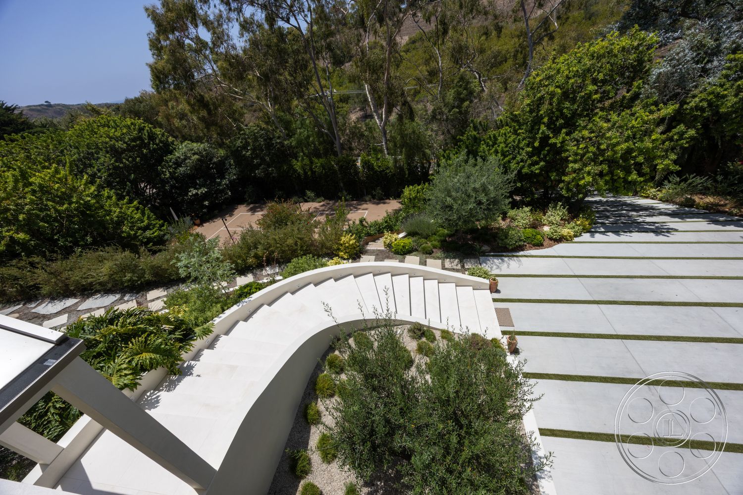 Americana 434 - exterior, outdoor seating area, landscaped garden area, minimalist architectural style, curved concrete steps, green shrubbery and trees, stone paver walkway, sloping hillside location, concrete retaining walls, concrete driveway with grass stripes