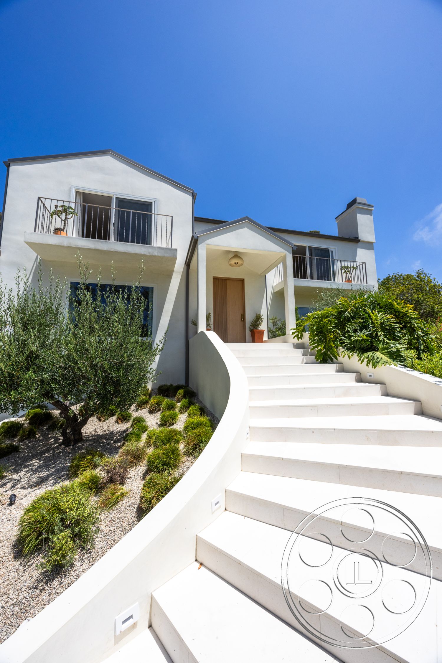Americana 434 - exterior, landscaped garden, white stucco walls, curved pathway, concrete steps, modern architectural style, green shrubbery, metal balcony railings, decorative potted plants, wooden front door, two-story house, gravel ground cover, gable roof