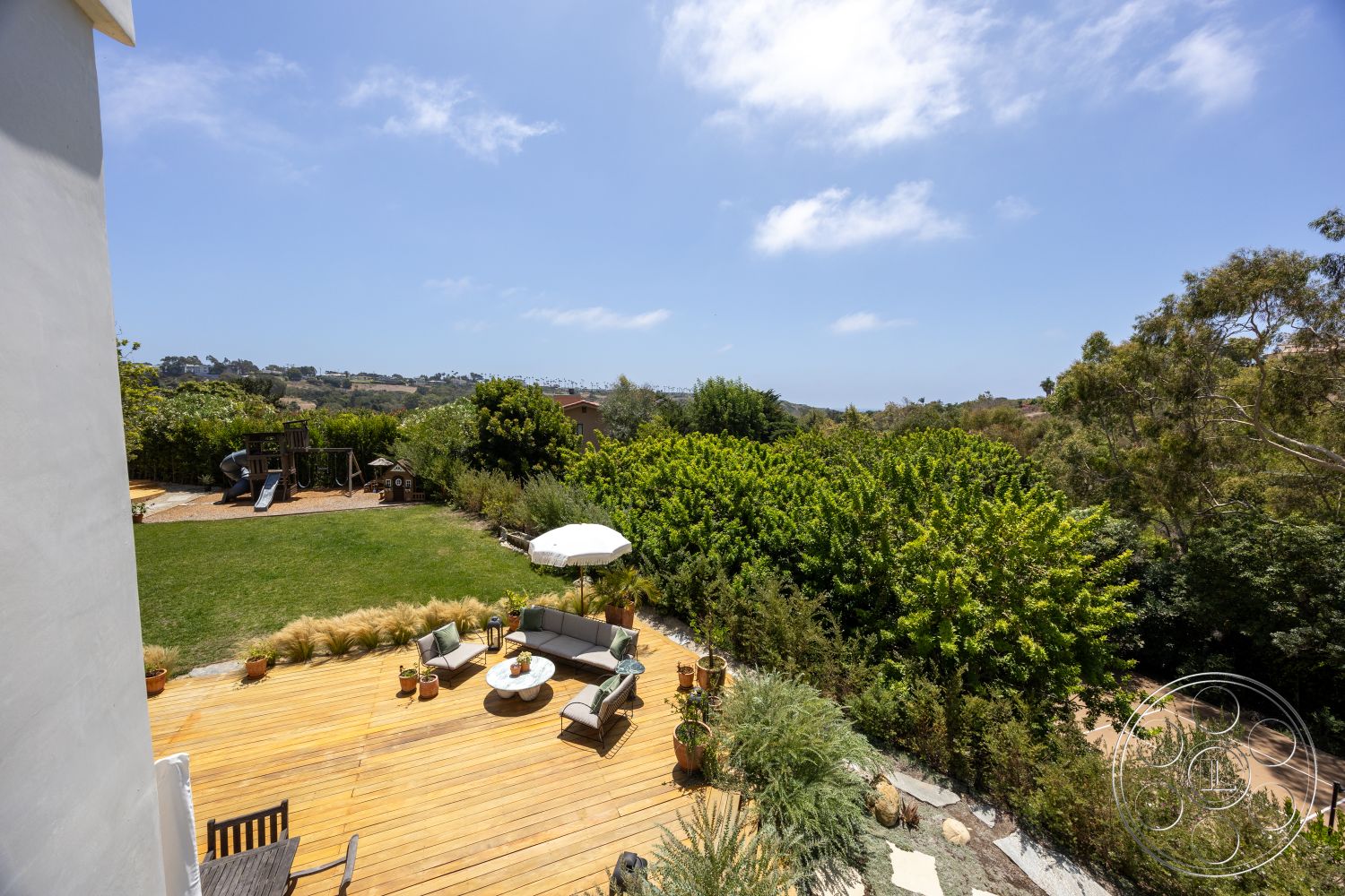 Americana 434 - exterior, landscaped garden, round coffee table, scenic hillside view, outdoor umbrella, lush green foliage, modern outdoor seating area, wood deck patio flooring, playground structure, open backyard