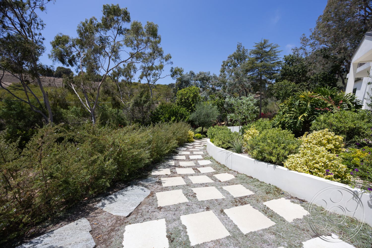 Americana 434 - exterior, natural vegetation, surrounding trees, stone pathway, landscape plants, trimmed hedges, sunny outdoor setting, rectangular stone pavers, white retaining wall