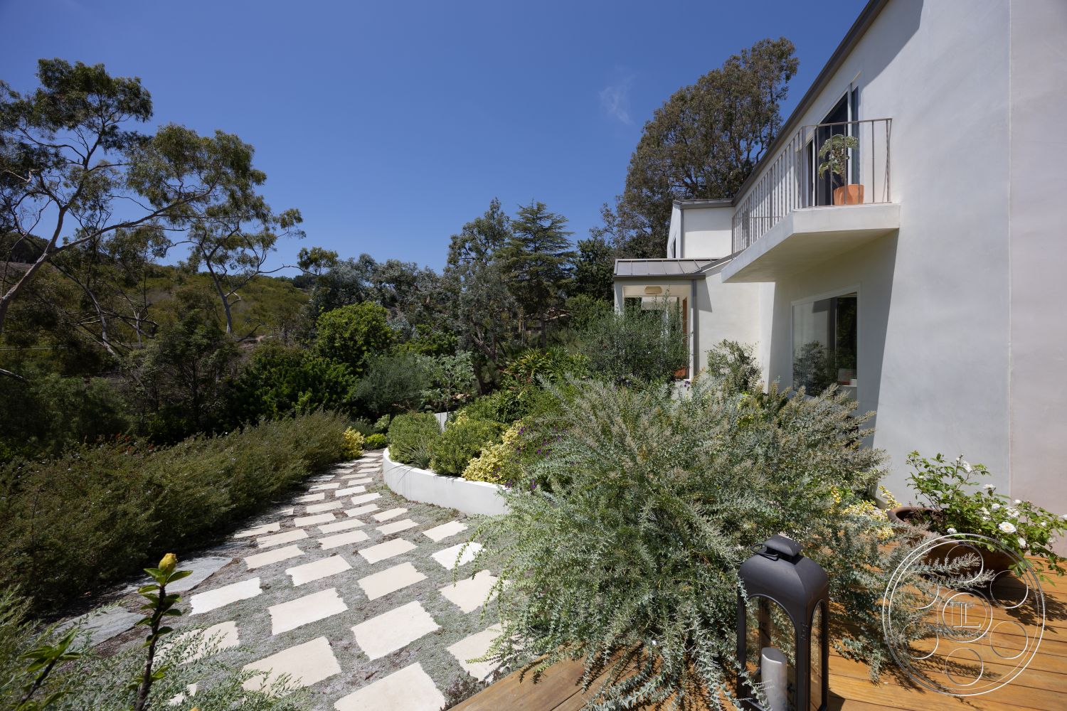 Americana 434 - exterior, landscaped garden, white stucco walls, concrete pathway, modern architectural style, balcony with railing, sunny outdoor area, hillside setting, wood deck flooring, square stone pavers, green plants and shrubs
