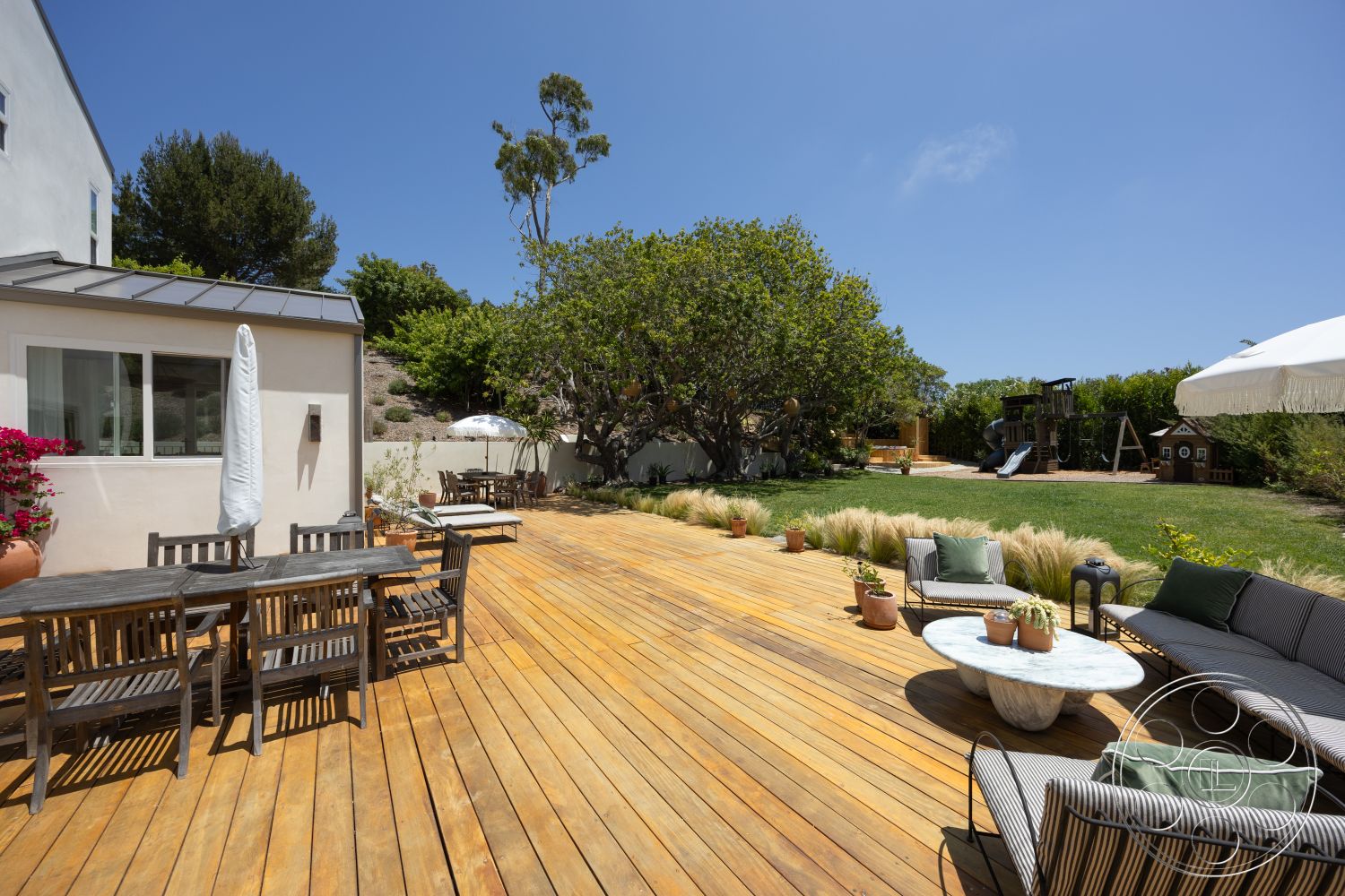 Americana 434 - exterior, potted plants, grassy lawn, blue sky, metal roof, outdoor entertaining area, landscaped garden area, wooden deck flooring, contemporary home facade, rectangular outdoor dining table, large shade tree, modern outdoor lounge seating, stainless steel outdoor umbrella, play area with swing set