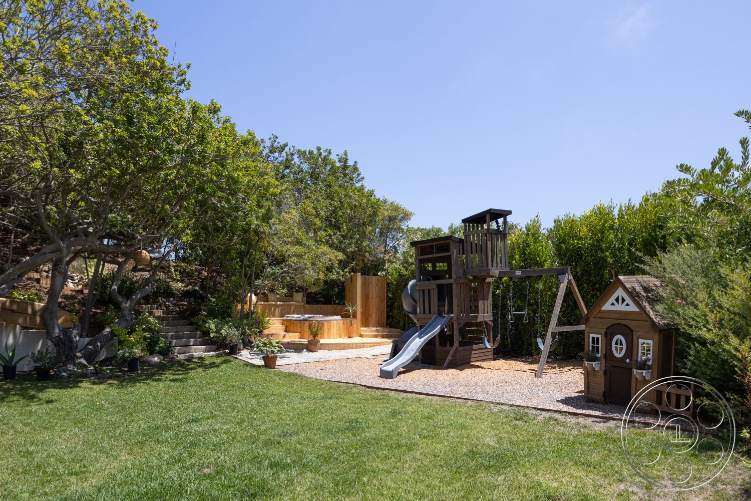 Americana 434 - exterior, outdoor seating area, wooden decking, surrounding trees, lush green lawn, wooden playhouse, swingset, landscaped garden beds, wood playground structure, sliding board, gravel play area, tiered stone steps