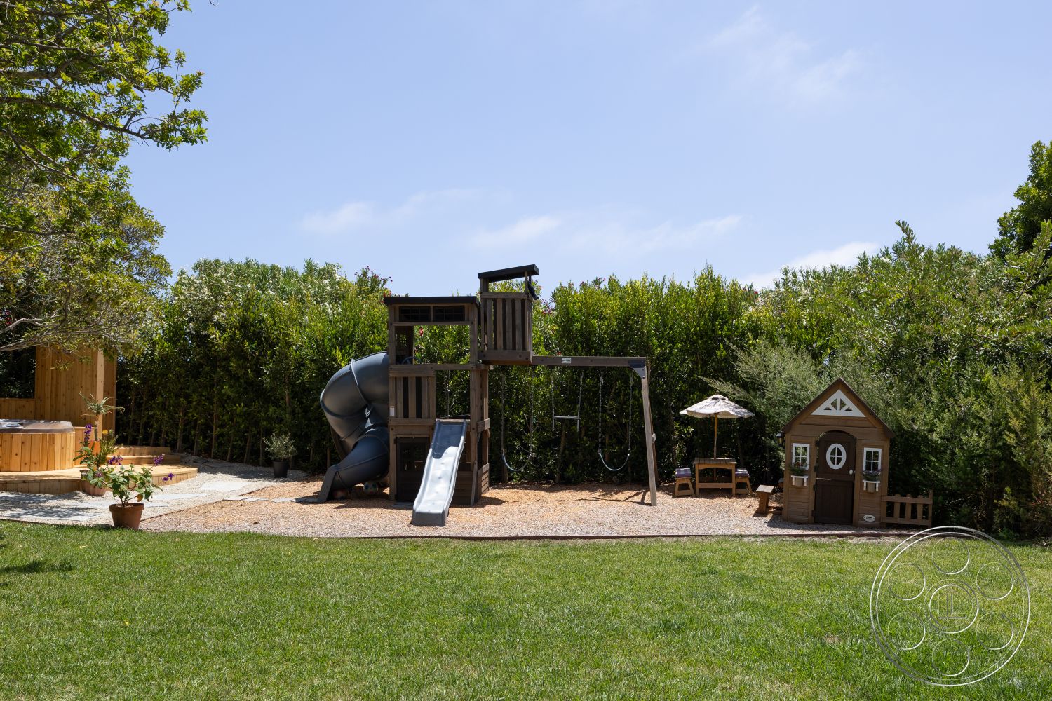 Americana 434 - exterior, wooden play structure, lush green grass lawn, surrounding hedge landscaping, gravel play area, metal slide, swing set with two swings, grey plastic slide, wooden playhouse with decorative trim, round wooden picnic table with umbrella, wooden hot tub structure