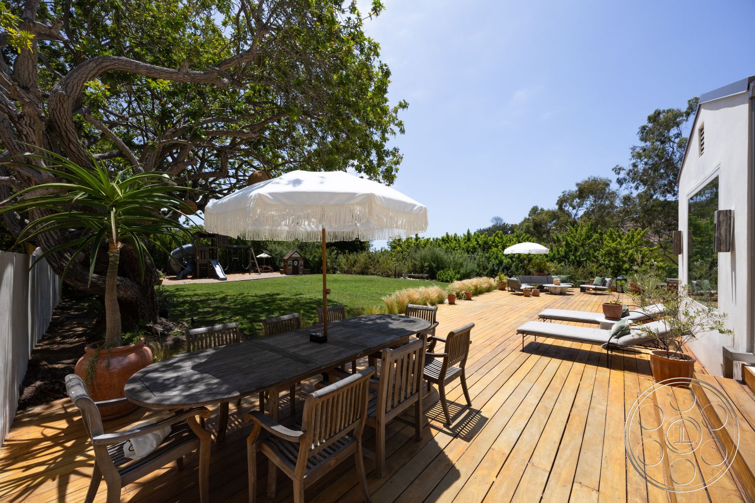 Americana 434 - natural light, exterior, wooden chairs, outdoor seating area, lush green lawn, sun loungers, rectangular dining table, smooth white walls, play structure, aluminum window frames, wood deck flooring, ornamental potted plants, modern home exterior, large patio umbrella, landscape shrubbery, large tree canopy, circular planter
