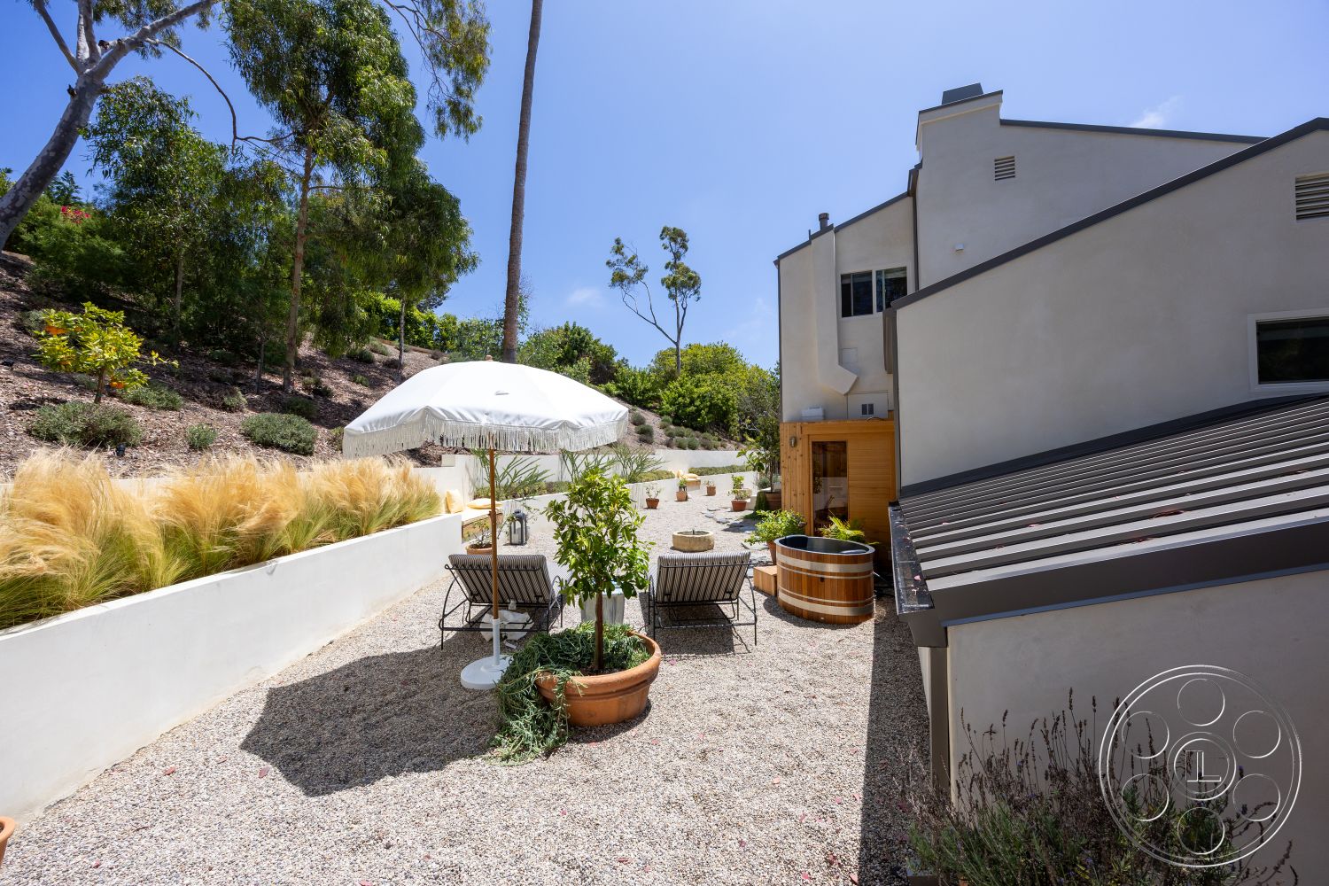 Americana 434 - exterior, outdoor dining area, planters with greenery, desert landscaping, stucco walls, curved garden beds, natural stone path, modern home, deck chairs, flat roof design, gravel patio flooring, urban hillside setting, wooden deck railing, white umbrella shade