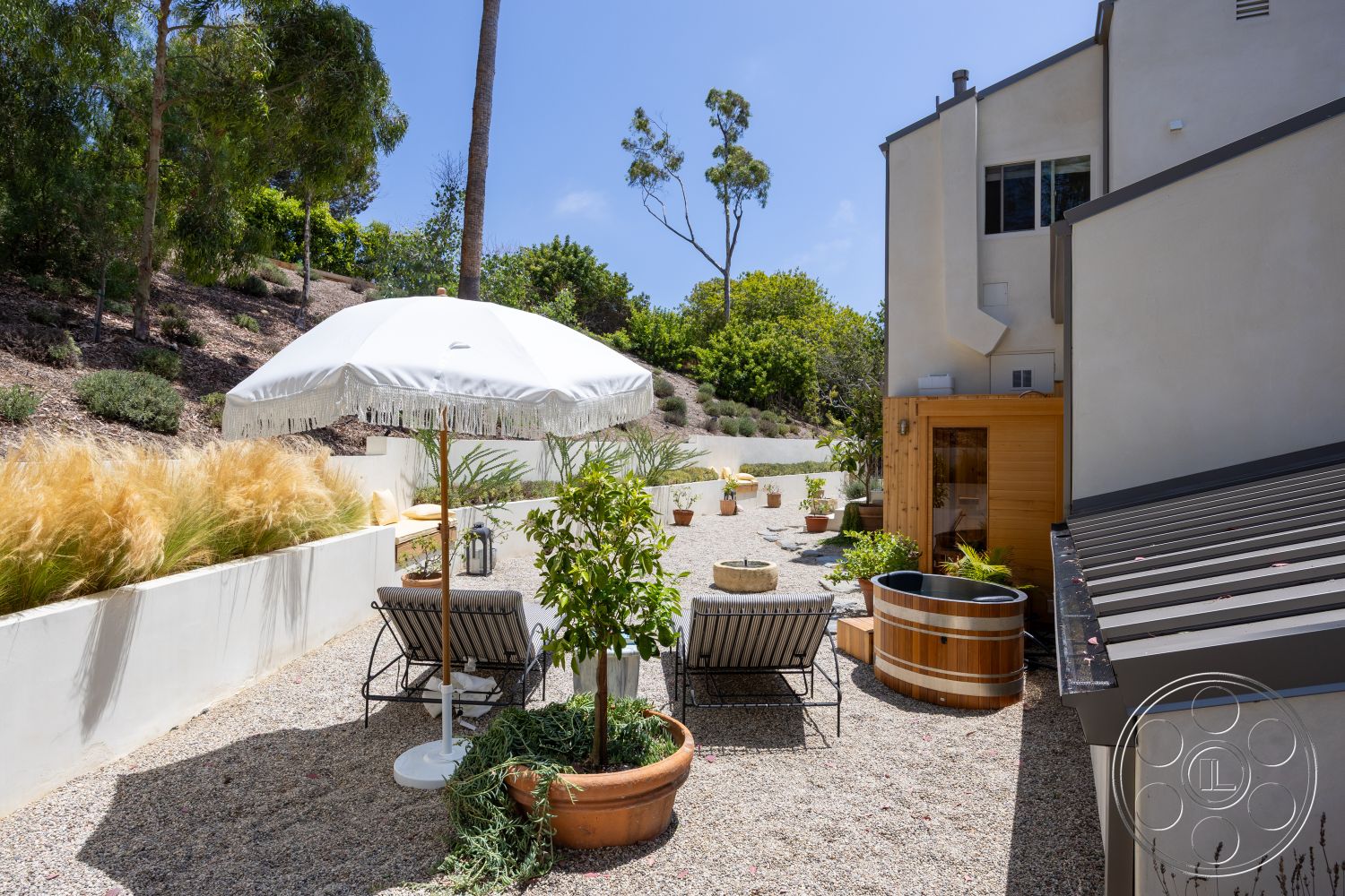 Americana 434 - exterior, black metal chairs, decorative lanterns, colorful potted plants, gravel patio flooring, sloped landscape setting, white concrete garden wall, wooden sauna exterior, modern metal roof, white parasol with fringe, tile balcony details, planters with lush greenery