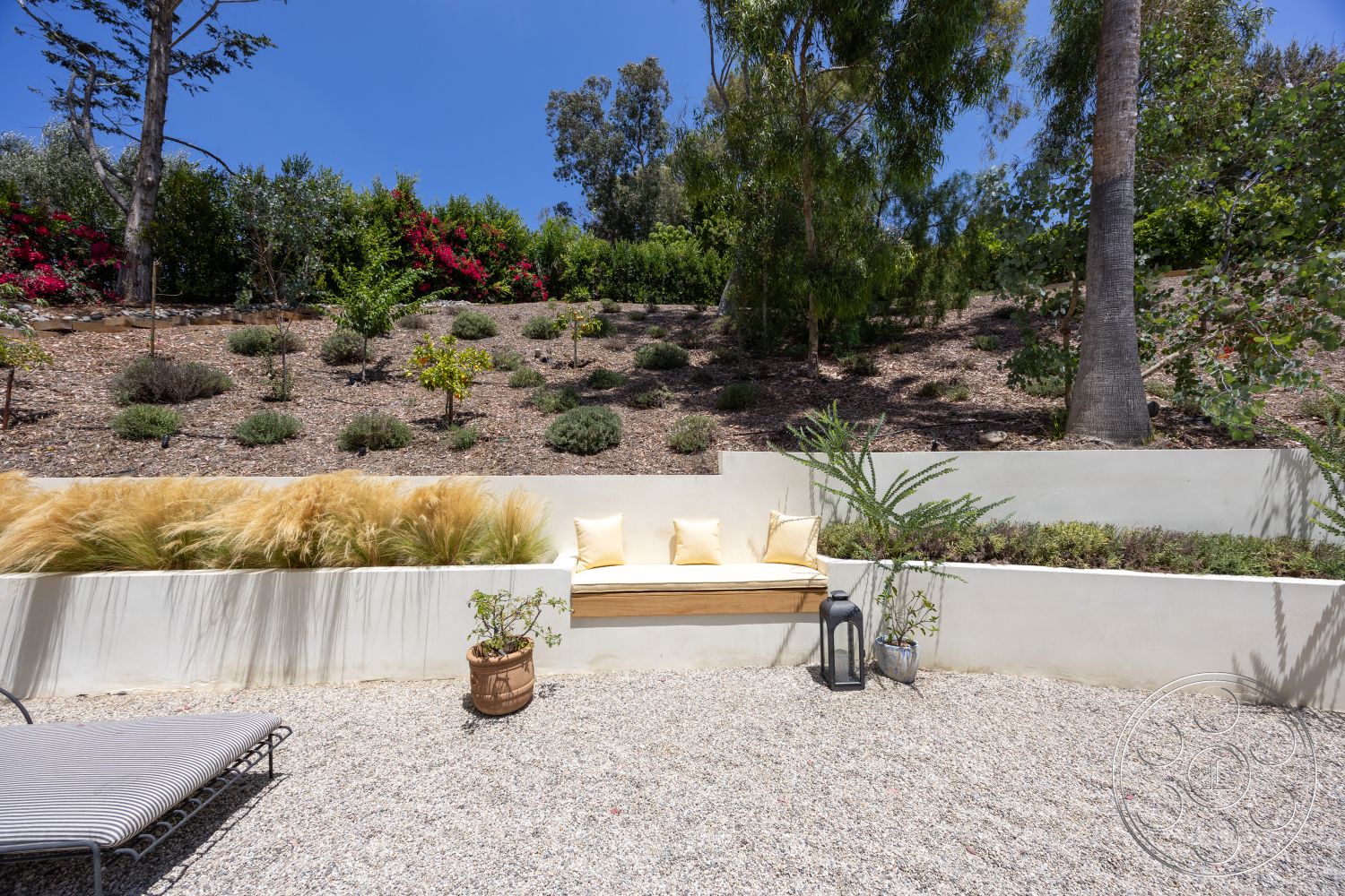 Americana 434 - exterior, potted plant, decorative lantern, gravel patio flooring, sunny outdoor seating area, landscaped hillside garden, natural stone ground cover, succulent plant arrangements, concrete bench with cushions, low-maintenance ornamental grasses, agave and shrub plantings, Mediterranean-style landscaping