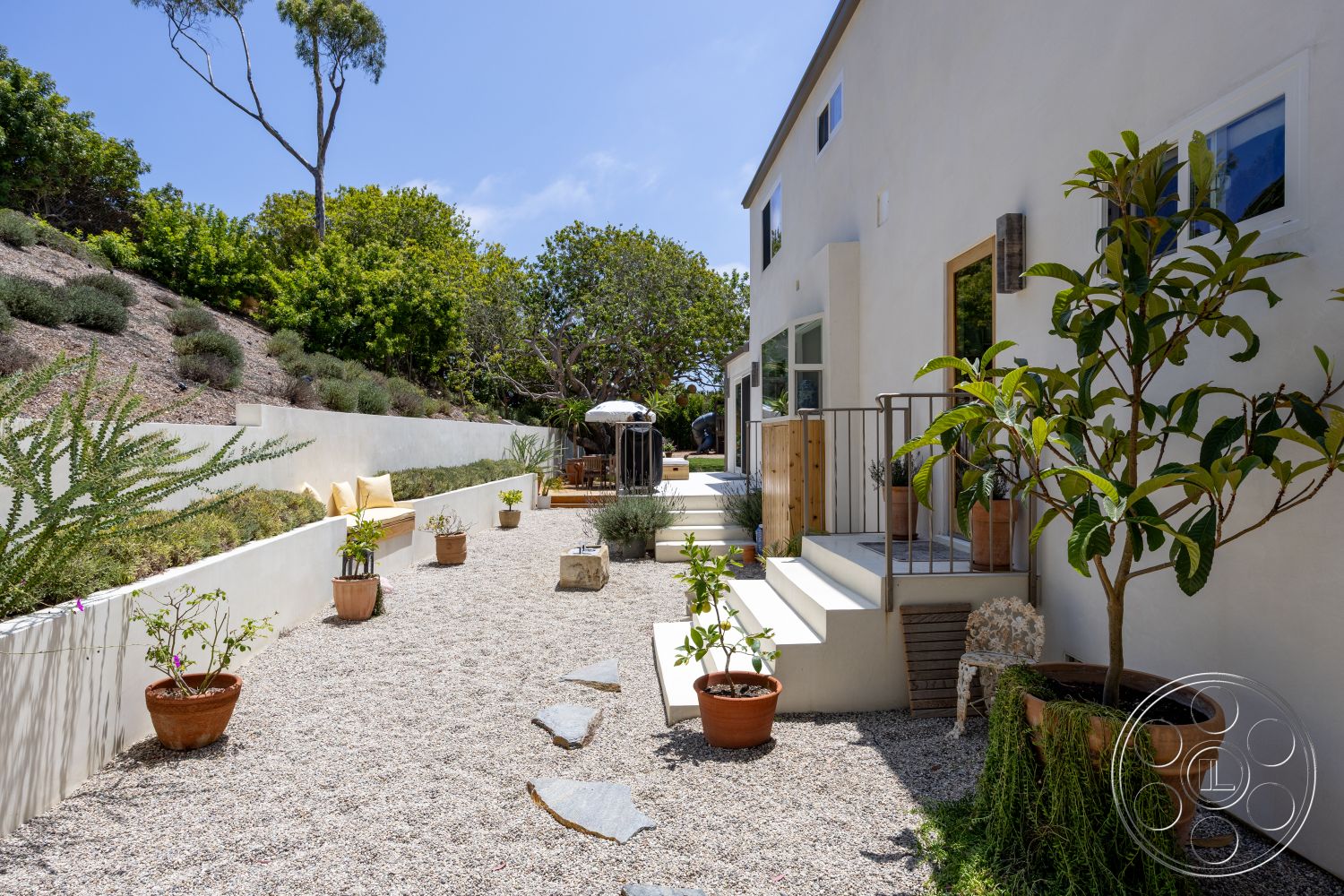 Americana 434 - exterior, natural stone accents, white stucco walls, modern architectural style, landscaped garden area, gravel patio flooring, open outdoor seating area, sunny urban setting, planter pots with greenery, large sliding glass windows, concrete step pathway, wood railing on staircase, lush hillside vegetation
