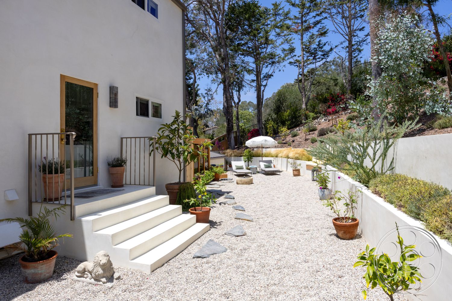 Americana 434 - exterior, landscaped garden, potted plants, outdoor seating area, metal railing, surrounding trees, concrete steps, stucco walls, sun umbrella, wood door, gravel patio flooring, decorative stone accents