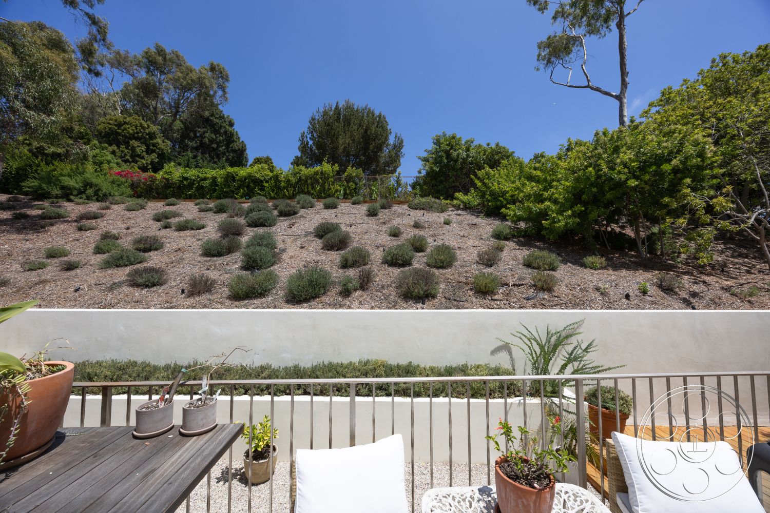 Americana 434 - exterior, lush greenery, potted plants, metal railing, clear blue sky, wooden outdoor furniture, concrete retaining wall, suburban backyard setting, gravel patio flooring, sloped landscape with low-maintenance plants