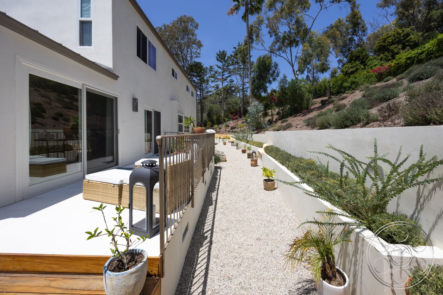 Americana 434 - exterior, glass sliding doors, wooden deck, gravel pathway, modern architectural style, landscaped hillside, tropical foliage, planter pots, concrete patio area, stucco house walls, concrete garden beds, parasitic plants