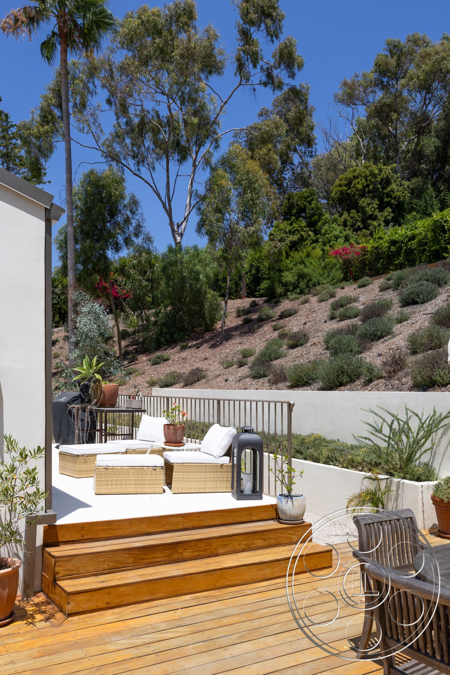 Americana 434 - exterior, wooden deck, tropical foliage, decorative potted plants, gravel ground cover, sunny outdoor setting, landscaped hillside garden, wicker patio furniture, railings with vertical metal slats, treaded wooden steps, concrete outdoor wall, flat seating area