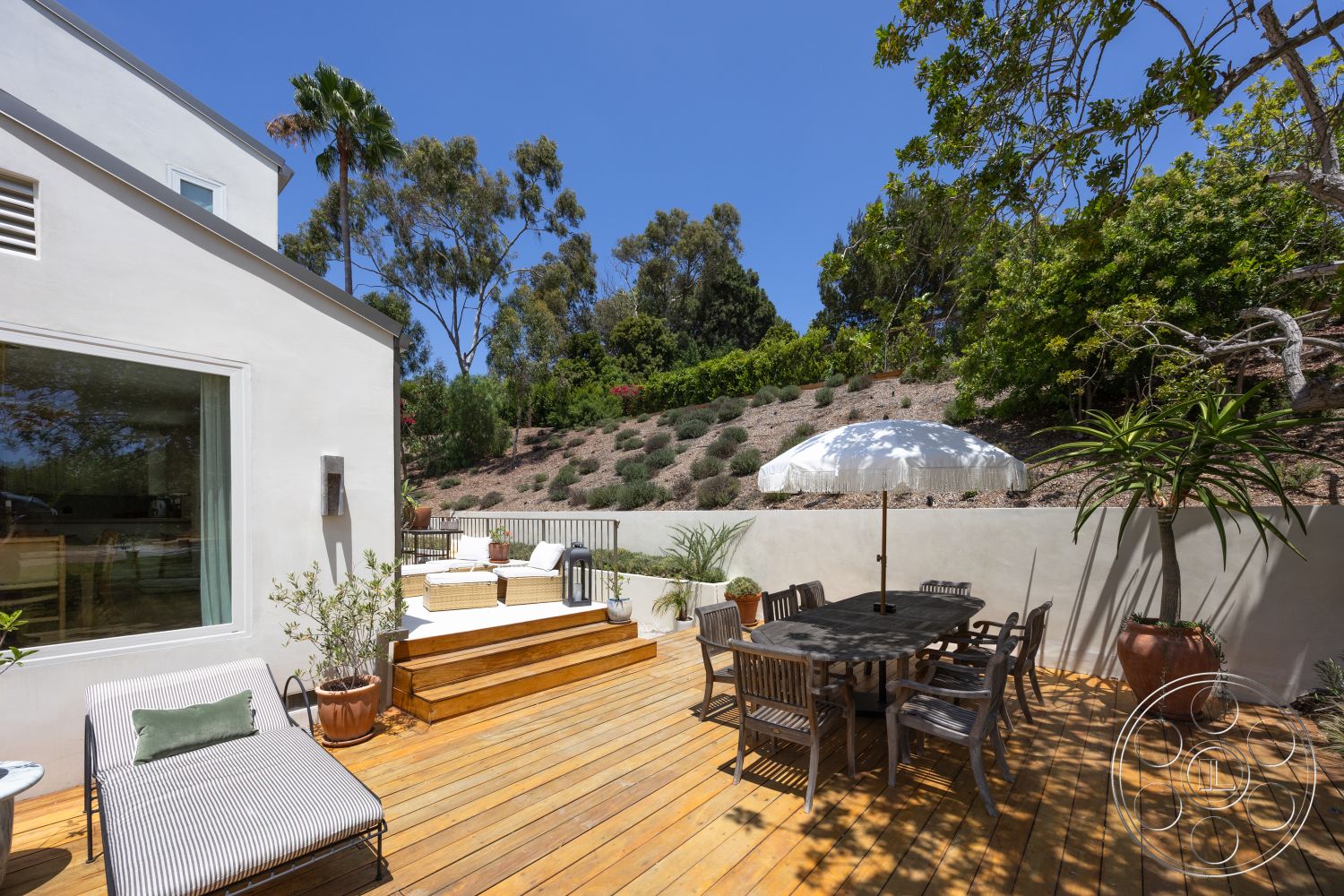 Americana 434 - exterior, rectangular dining table, wooden lounge chairs, wooden dining chairs, metal patio umbrella, wood deck patio flooring, sunny outdoor environment, landscaped hillside setting, plaster exterior walls, succulent plants in pots, large windows with light green curtains, dining area with outdoor seating
