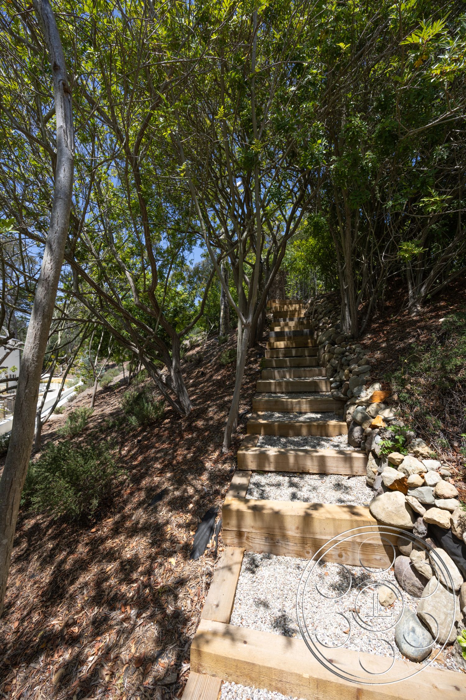 Americana 434 - exterior, landscaped hillside, surrounded by trees, natural stone retaining wall, forest setting, wooden outdoor stairs, gravel path surface, steps with wooden risers