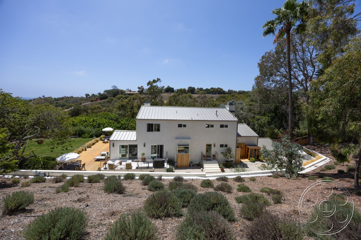 Americana 434 - large windows, exterior, white stucco walls, outdoor seating area, surrounding trees, metal roof, hardscaped pathway, hilltop location, rectangular patio layout, wooden deck patio, landscape garden setting, horizontal fence panels