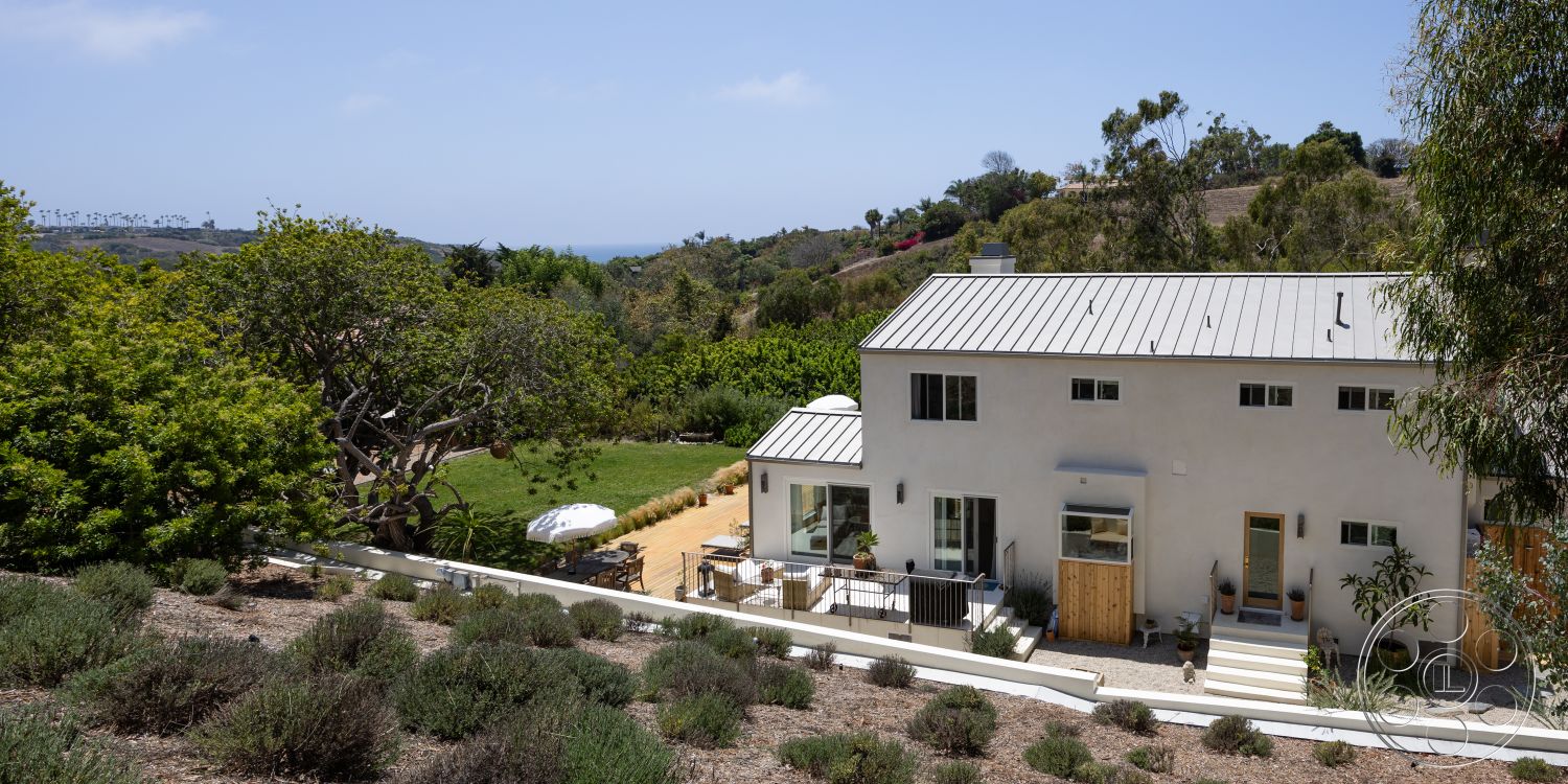Americana 434 - exterior, landscaped garden, white stucco walls, ocean view, metal roof, modern farmhouse style, wooden front door, outdoor umbrella, tall glass windows, minimalist architectural details, rectangular patio, sloped hillside, natural setting with greenery, lavender garden beds, wooden deck with outdoor seating, seating area with table and chairs