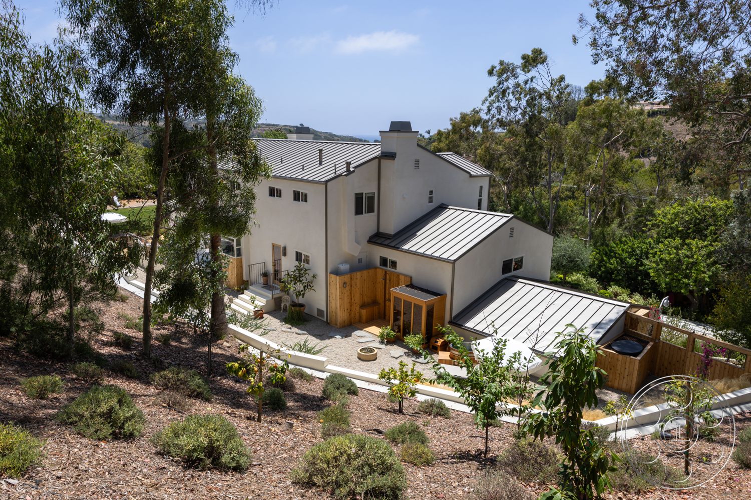 Americana 434 - exterior, white stucco walls, modern architectural style, metal roof, pathway leading to entrance, landscaped garden setting, wooden deck area, outdoor seating arrangement, sloped roof shape, raised flower beds, stone patio surface, eucalyptus trees surrounding