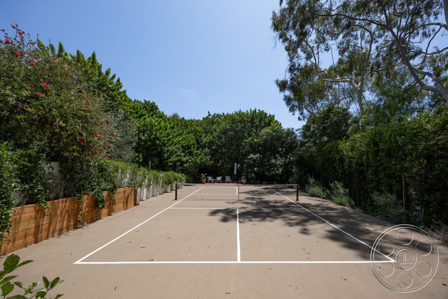 Americana 434 - exterior, landscaped garden, lush greenery, outdoor seating area, sunny outdoor environment, wooden privacy fencing, sand-based tennis court, white court markings