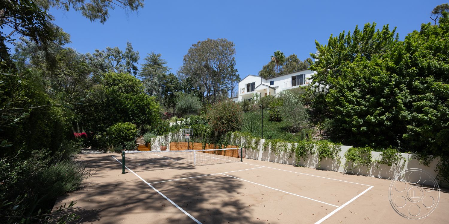 Americana 434 - exterior, surrounding trees, sunny outdoor setting, white painted walls, lush greenery landscaping, sloped hillside location, sand tennis court, wooden tennis net posts, concrete court boundary lines, modern house with large windows, mixed vegetation types, raised wooden planters