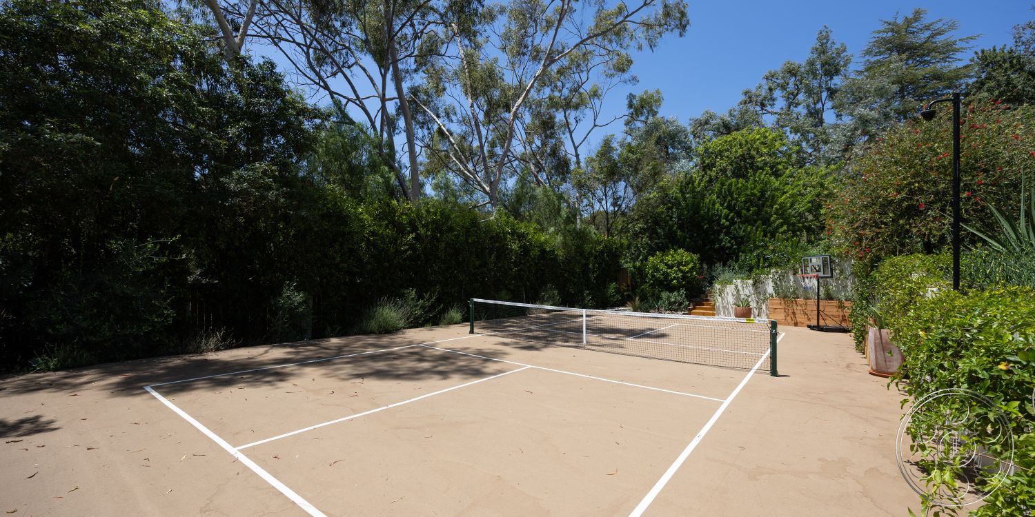 Americana 434 - exterior, surrounding greenery, wooden planter boxes, basketball hoop, bush landscaping, sunny outdoor setting, tennis court lines, sand court surface, dense trees