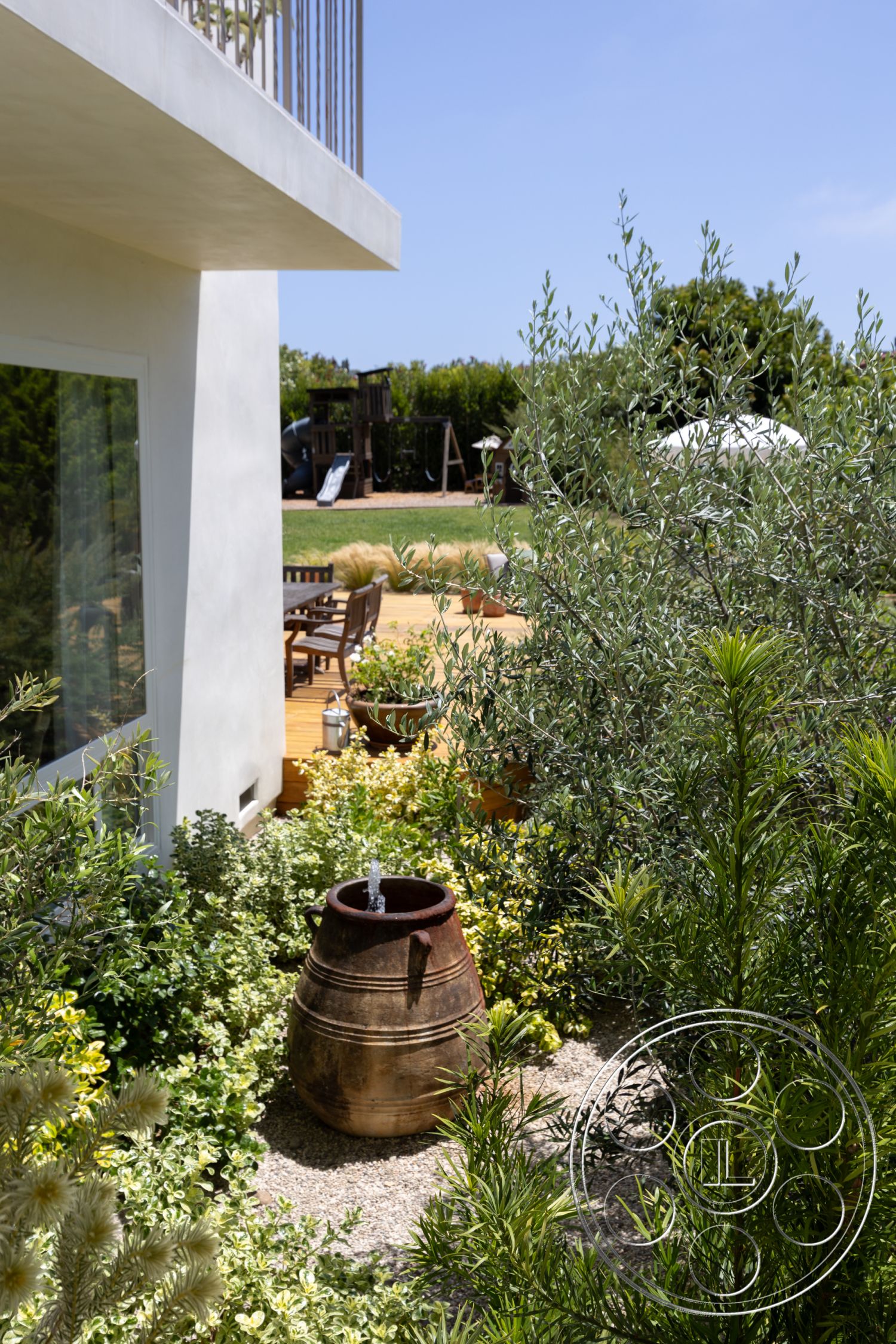 Americana 434 - exterior, modern architectural style, stucco wall finish, gravel garden path, landscaped garden with native plants, sunny backyard setting, wooden outdoor dining furniture, parquet wood patio flooring, ceramic garden pot, clay water feature