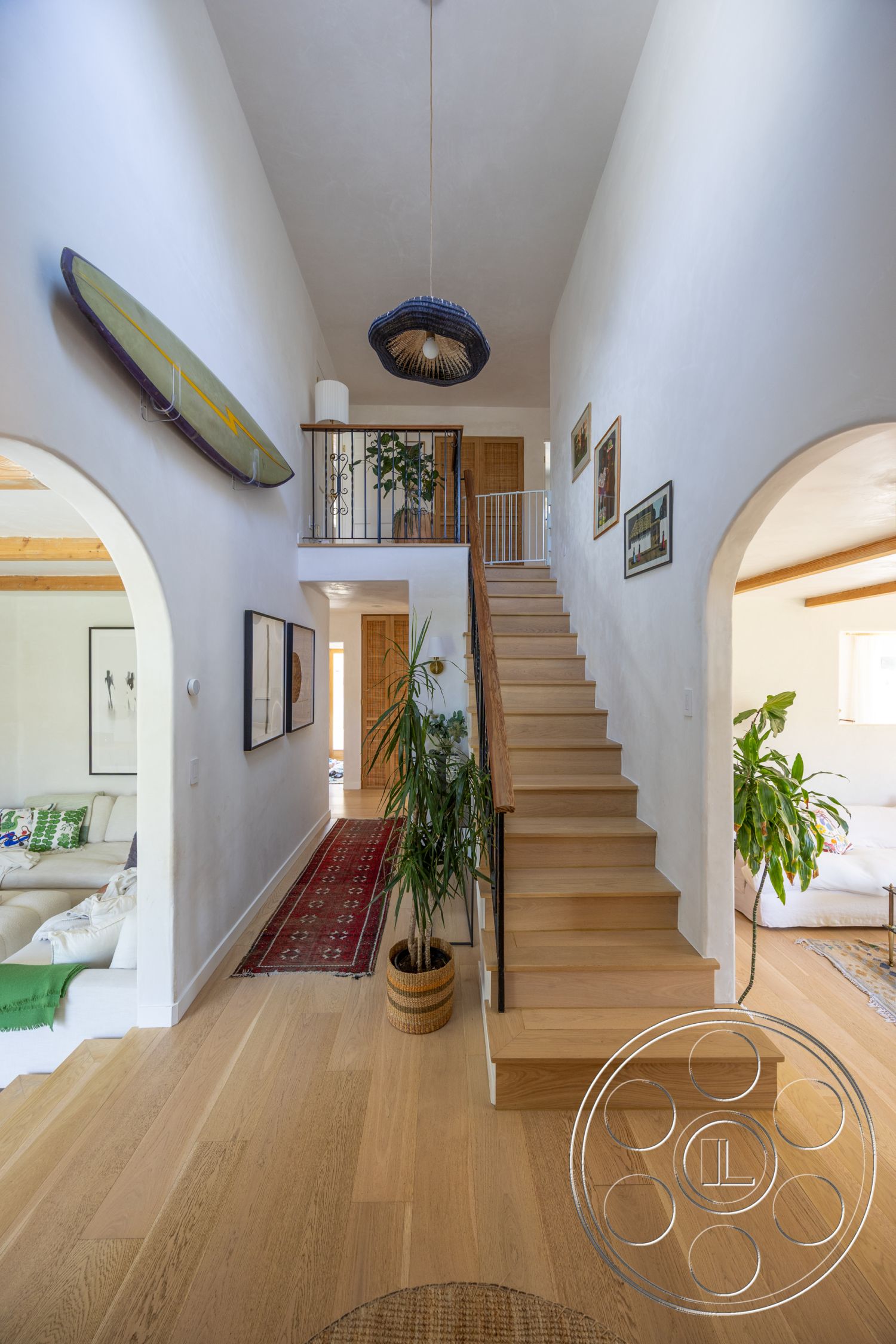 Americana 434 - neutral color palette, textured area rug, decorative wall art, arched doorways, white plaster walls, woven pendant light fixture, indoor potted plants, black metal stair railings, natural wood ceiling beams, light oak hardwood flooring, open staircase with wooden treads, woven basket planter, cozy entrance hallway