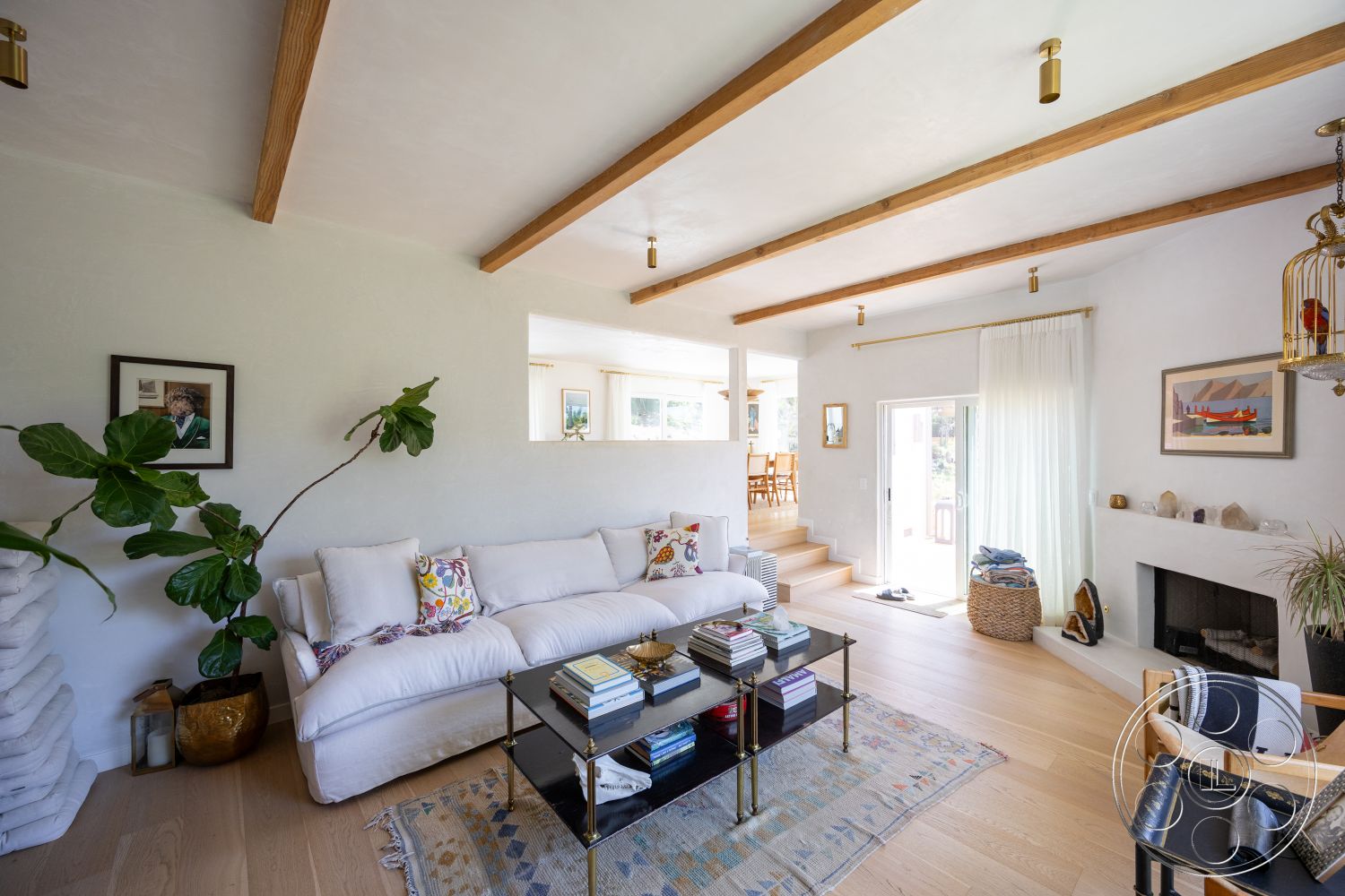 Americana 434 - decorative area rug, floor-to-ceiling windows, green potted plant, light-colored curtains, exposed wood ceiling beams, hardwood living room floors, woven basket for storage, stone fireplace surround, white textured plaster walls, modern sofa with decorative cushions, glass coffee table with books, black framed wall art, staircase leading to dining area