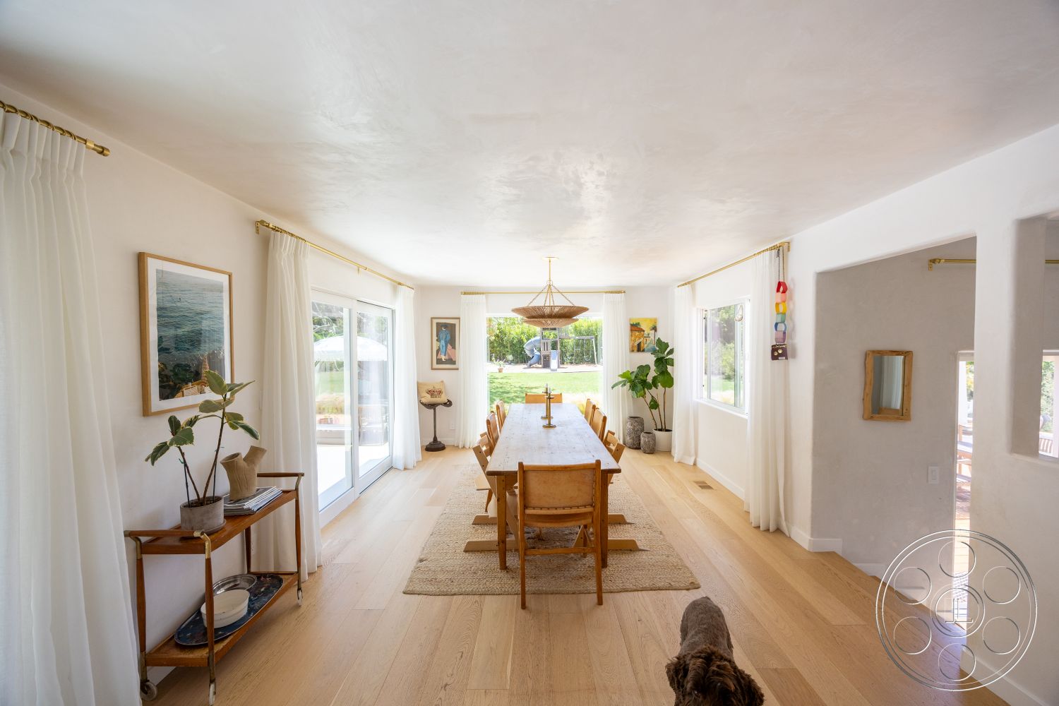Americana 434 - minimalist decor, neutral color palette, natural light, open floor plan, artwork on walls, seamless indoor-outdoor transition, floor-to-ceiling windows, green indoor plants, textured ceiling, rug under dining table, contemporary light fixture, light oak hardwood flooring, white painted plaster walls, dining area with large wooden dining table, curtains with gold hardware, stylish wooden side table