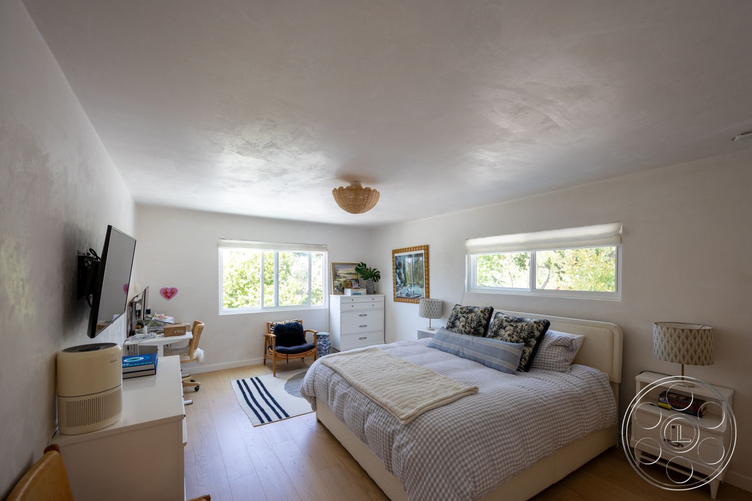 Americana 434 - decorative plant, striped area rug, framed wall art, plaster painted walls, modern ceiling light fixture, cozy reading chair, light wood bedroom floors, queen-sized bed with patterned bedding, glass windows with white trim, white nightstands with lamps, desks with white surfaces, family-friendly bedroom