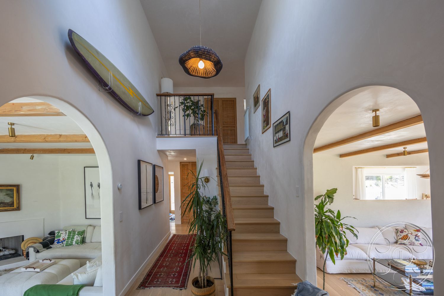 Americana 434 - light hardwood flooring, wooden staircase, textured plaster ceiling, woven area rug, arched doorways, white plaster walls, woven pendant lighting, exposed wood ceiling beams, open living space layout, modern sofa, decorative indoor plants, metal railing details, framed artwork on walls, light wood stair treads