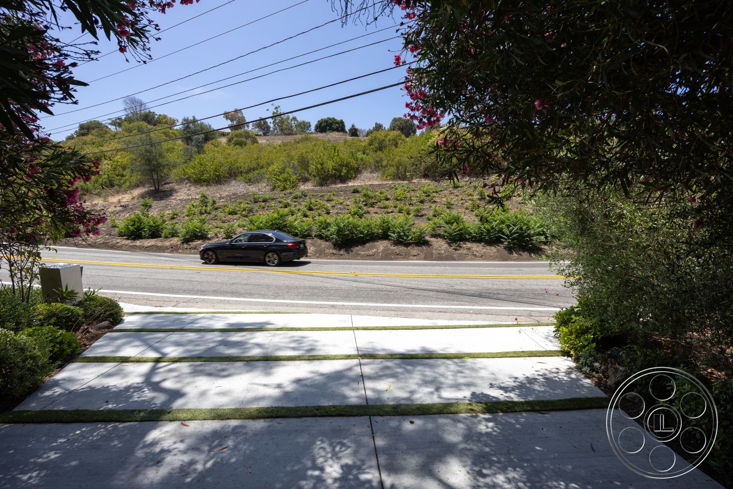Americana 434 - exterior, paved road, clear sky, flowering shrubs, concrete driveway, mountain background, landscaped garden beds, striped grass sections, dirt hillside