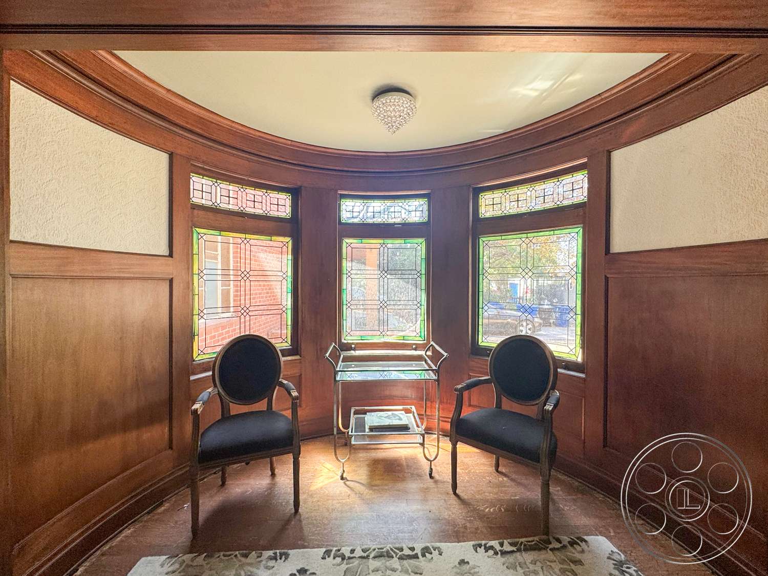 The Rosenheim Mansion - hardwood flooring, crystal chandelier, stained glass windows, dark wood paneling, patterned area rug, curved bay window, round ceiling detail, antique accent chairs, glass top side table
