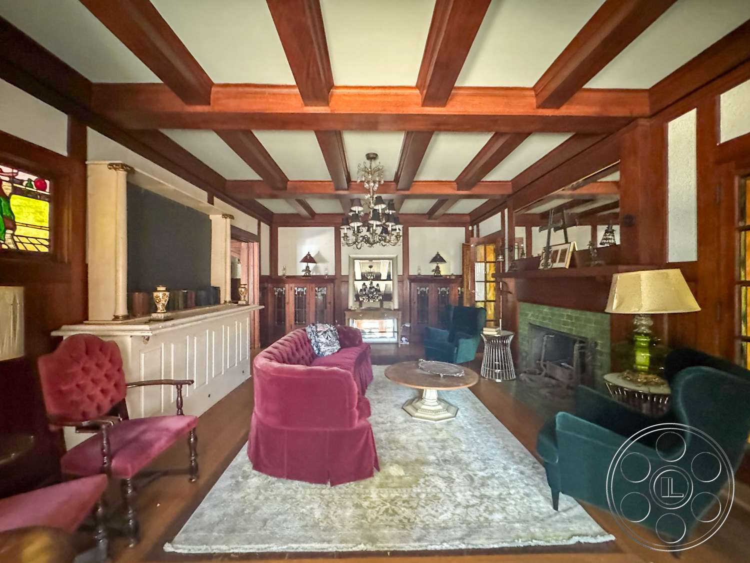 The Rosenheim Mansion - coffered ceiling, white wainscoting, circular coffee table, stained glass windows, vintage armchair, dark wood paneling, dark wood ceiling beams, traditional chandelier, hardwood living room floors, green velvet armchair, intricate wall moldings, plush burgundy sofa, green tile fireplace, art deco style lamp, period-style decor