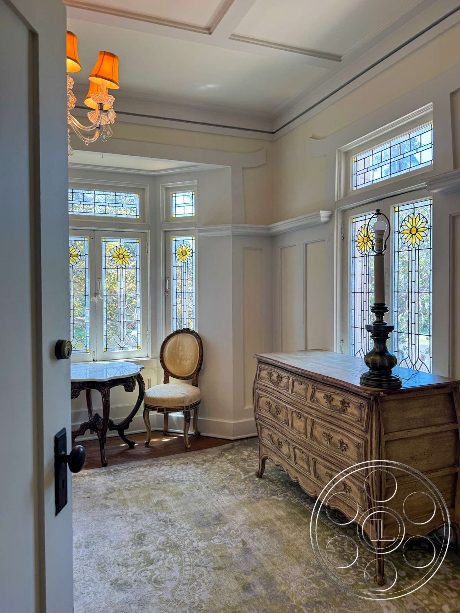 The Rosenheim Mansion - decorative area rug, wooden flooring, natural light from large windows, stained glass windows, geometric ceiling design, vintage chandelier, cream painted walls, ornate crown molding, elegant room layout, carved wooden dresser, antique upholstered chair, side table with decorative legs